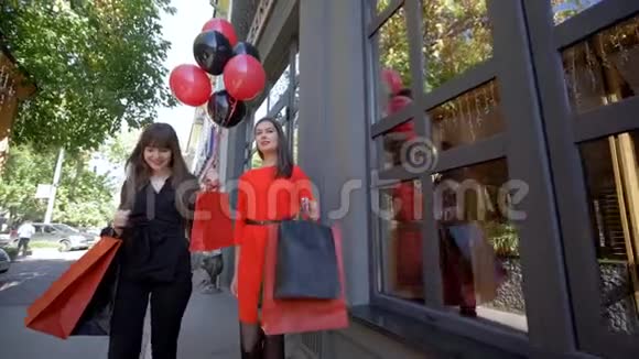 季节性销售快乐的时尚女孩带着袋子在购物时交流走过黑色精品店的窗户视频的预览图