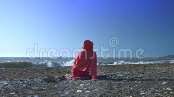 后景有人穿着红色运动衫和引擎盖在暴风雨中坐在海滩上看着大海视频的预览图