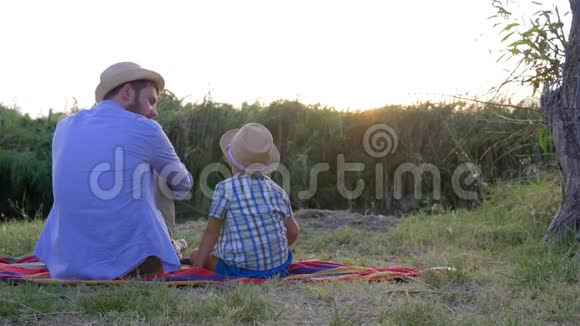 童年快乐小男孩和爸爸戴着草帽坐在露天的毯子上享受日落视频的预览图