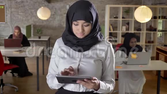年轻的穆斯林妇女戴着头巾使用平板电脑看着镜头微笑着商业妇女在现代办公室的笔记本电脑上打字视频的预览图