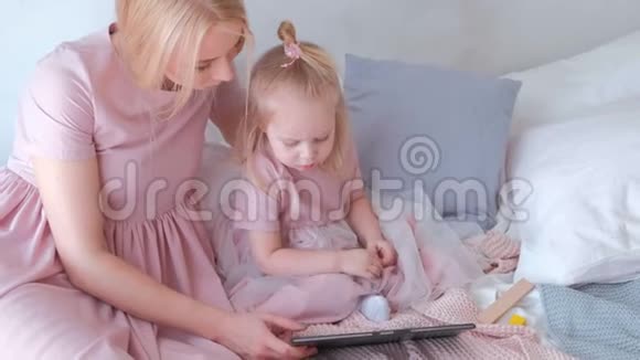年轻迷人的金发女郎和她的小女儿穿着粉红色的连衣裙坐在平板电脑里看着什么东西视频的预览图