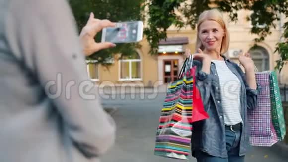 快乐购物狂摆姿势智能手机相机拿着购物袋在户外视频的预览图