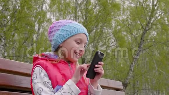 画中的少年女孩在绿树背景的春天公园里用手机视频的预览图