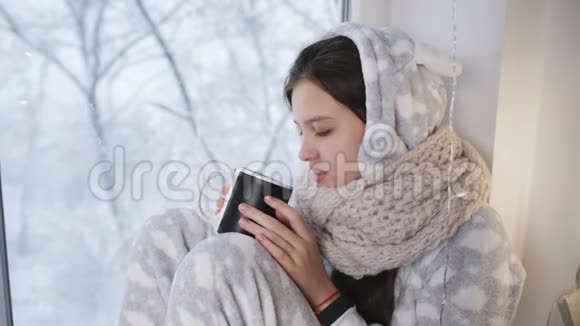生病悲伤的女孩少年穿着温暖的睡衣坐在窗前喝着一杯热茶视频的预览图