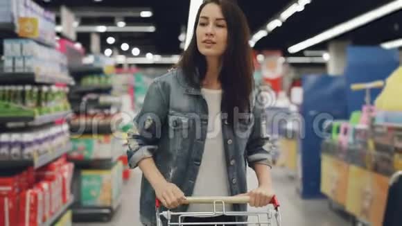 漂亮的女人开着购物车穿过超市的食品部门四处张望漂亮女孩视频的预览图