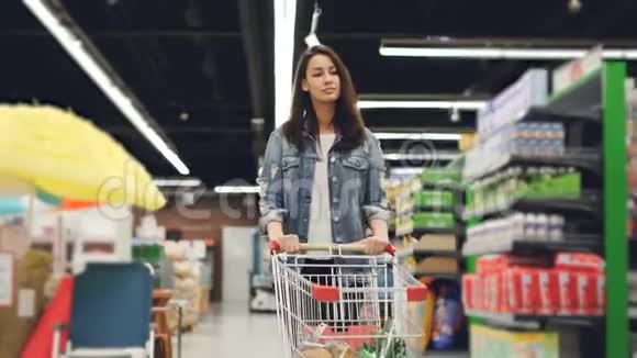 多莉拍了一张漂亮的年轻女士的照片她带着购物车沿着一排排的货架走着带着产品环顾四周视频的预览图