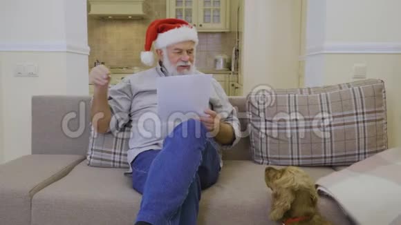戴着圣诞老人帽的老人在家沙发上读心愿单视频的预览图