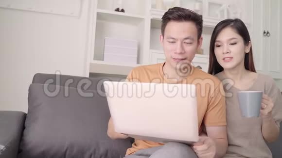 亚洲夫妇使用笔记本电脑在家里的客厅里喝着温暖的咖啡甜蜜的夫妇享受的时刻视频的预览图