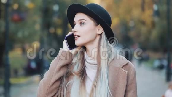 一位年轻的美女站在秋天的公园里站在一片黄叶背景下用手机说话视频的预览图