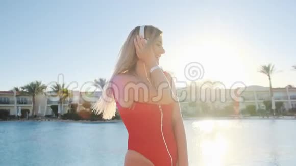 美丽的女士穿着红色泳衣戴着耳机在清澈的蓝色游泳池附近跳舞玩得开心听音乐视频的预览图