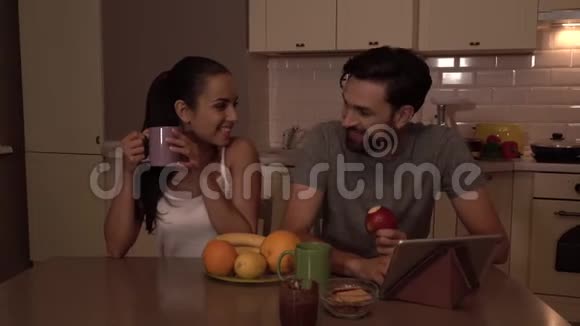 幸福的小家庭成员坐在桌边女孩从杯子里喝他吃苹果有平板电脑和盒装视频的预览图