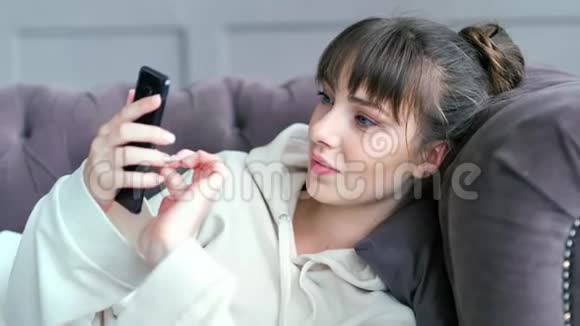 年轻迷人的女人躺在沙发上用智能手机聊天的肖像视频的预览图