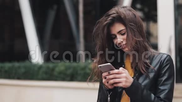 漂亮的年轻女人穿着皮夹克站在市中心环顾四周时用她的手机城市视频的预览图
