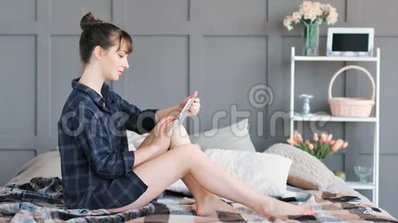在坐在床上的社交网络中微笑着用平板电脑写信息聊天的年轻女子视频的预览图