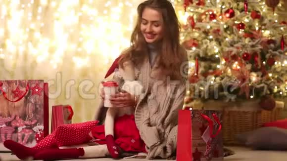 带着格子呢和圣诞老人的年轻女人坐在圣诞树和礼品包旁边微笑着喝咖啡视频的预览图