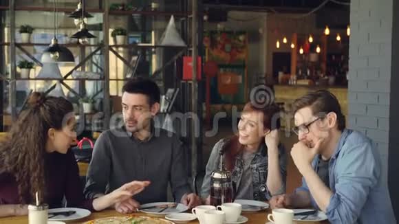 快乐的年轻朋友正在现代咖啡馆里坐在餐桌旁交谈和手势分享新闻大比萨杯子和视频的预览图