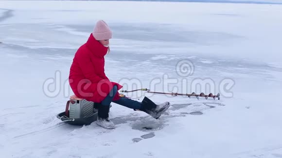 穿着红色夹克和毛毡靴的年轻美女坐在冰上借助冬天的鱼竿在洞里钓鱼视频的预览图