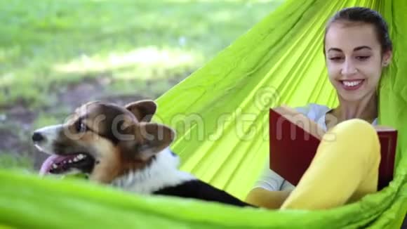 迷人的年轻女子与可爱的威尔士科尔吉狗躺在明亮的绿色吊床在公园和阅读一本书视频的预览图