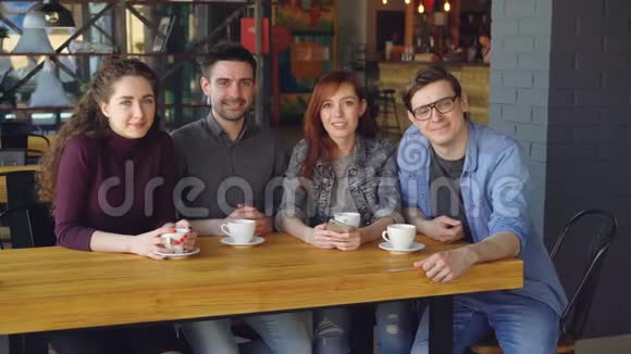 四个穿休闲服装的年轻人朋友坐在桌子旁在宽敞的咖啡馆里喝着茶杯看着视频的预览图