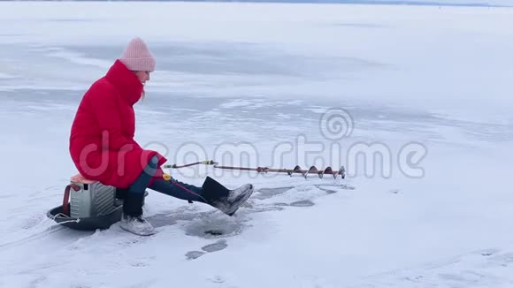 穿着红色夹克和毛毡靴的年轻美女坐在冰上借助冬天的鱼竿在洞里钓鱼视频的预览图