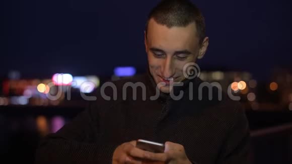 男人一边微笑一边自拍或在夜间灯光下在户外观看照片夜间使用智能手机短信视频的预览图
