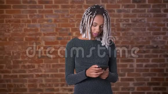年轻的非洲女孩戴着可怕的锁用智能手机看着镜头微笑着背景是砖墙视频的预览图