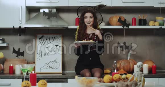 微笑着美丽的亚洲女孩蒙面在厨房的女巫坐在厨房的桌子上玩一些零食视频的预览图