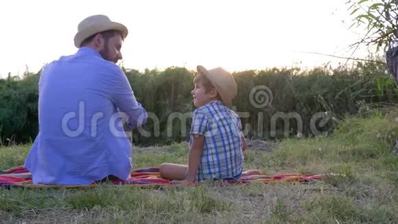 父子关系爸爸带着戴草帽的小男孩坐在露天的毯子上享受日落视频的预览图