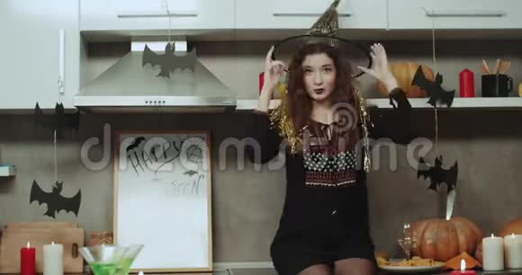 一个卷发女巫在镜头前跳舞和移动魅力她坐在厨房的桌子上视频的预览图