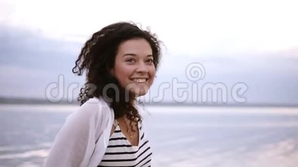 美丽的年轻卷曲的黑发女子赤脚在海洋或湖泊中行走快乐微笑害羞的女性穿着条纹T恤视频的预览图
