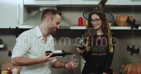 一对微笑的年轻夫妇在万圣节派对上在厨房的令人惊叹的装饰中喝着酒杯中的红酒视频的预览图