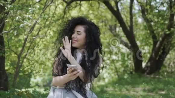 一个漂亮的黑发女人在花园里和一个人打招呼时用她的电话视频的预览图
