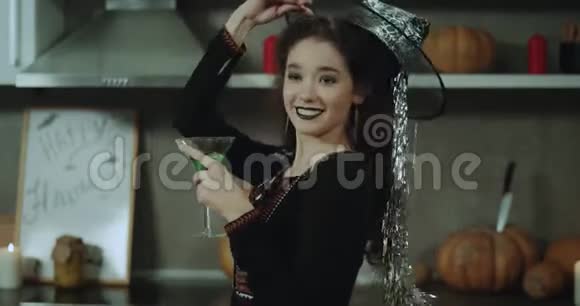 在厨房里一位戴着女巫面具的贴身卷曲的年轻女士准备参加万圣节派对喝一杯绿色鸡尾酒视频的预览图