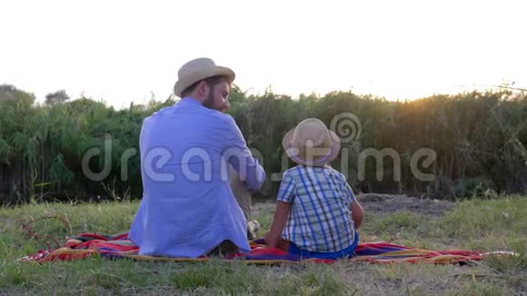 可爱的父亲和戴草帽的可爱的儿子一起在大自然中度过时光坐在露天的毯子上视频的预览图