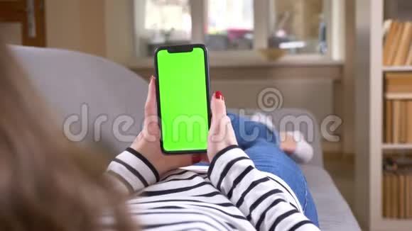 特写后景拍摄女性使用手机与绿色屏幕保持它垂直躺在沙发在室内视频的预览图