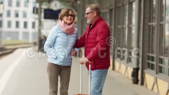 成熟的夫妇带着手提箱在火车站台上笑着拥抱旅行退休人员一男一女穿着视频的预览图