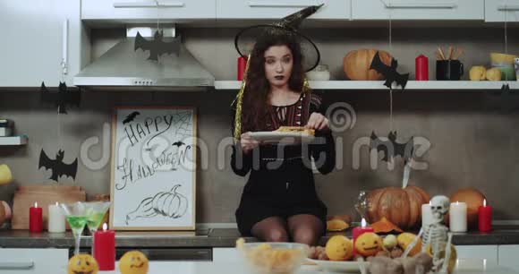 一个年轻的蒙面女巫坐在厨房的桌子上拿着一个小吃盘在镜头前玩视频的预览图