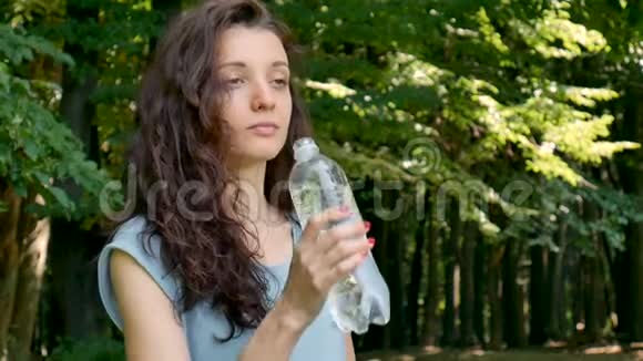 户外画的年轻不伦特女孩的卷发是饮用矿泉水的绿树背景视频的预览图