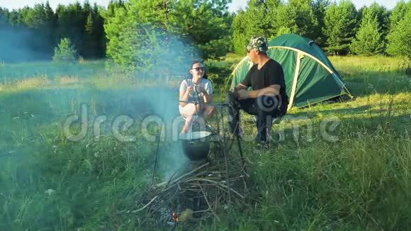 男人和女人的游客坐在帐篷附近的篝火旁看着锅挂在燃烧的火堆上视频的预览图