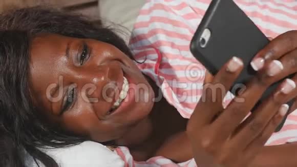 美丽的美国黑人女士穿着睡衣躺着轻松的微笑愉快的新鲜的使用互联网手机网络视频的预览图