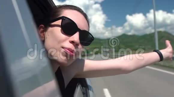 戴着太阳镜的快乐女孩斜倚在车窗外享受着在山里的旅行年轻女子望着窗外视频的预览图
