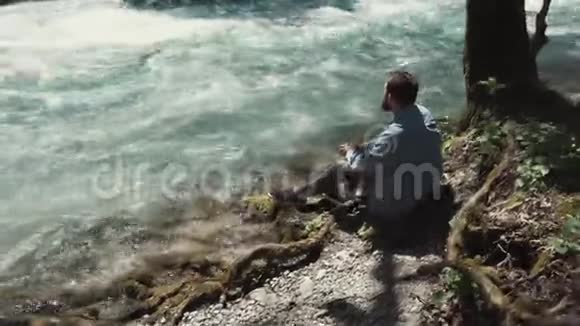 一个年轻人坐在河快流附近看着流水视频的预览图