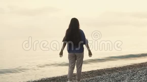 迷人的黑发女人站在夕阳下的鹅卵石海滩上双手向上举起视频的预览图