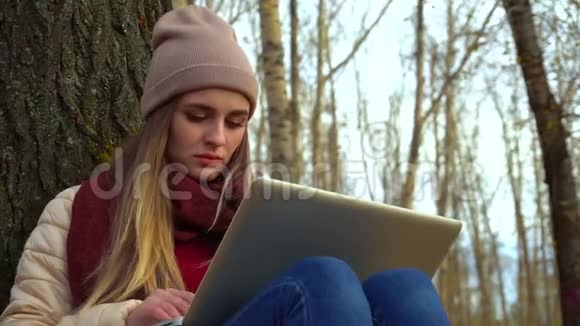迷人的年轻女孩在秋天公园的树下坐在地上在笔记本电脑里工作快关门视频的预览图