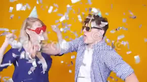 节日和生日的概念年轻的幸福夫妇戴着橙色背景的帽子和五彩纸屑跳舞视频的预览图