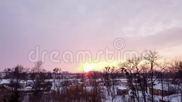 随着时间的推移冬天的日落笼罩着白雪覆盖的城市黑暗的光秃秃的树木轮廓房屋的屋顶橙色的太阳视频的预览图