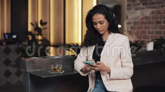 漂亮的女人使用智能手机坐在咖啡馆或同事办公室喝茶成功的肖像视频的预览图
