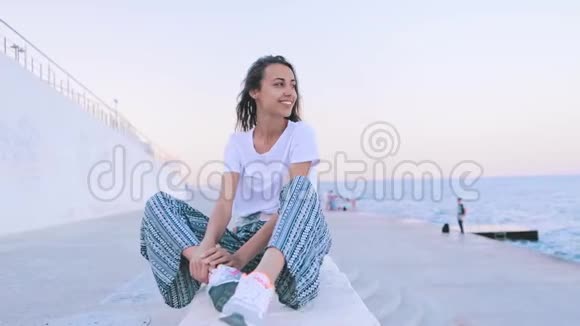 黎明时分美丽的年轻女子穿着轻盈的夏装坐在码头上年轻的白种人女性正在观看视频的预览图