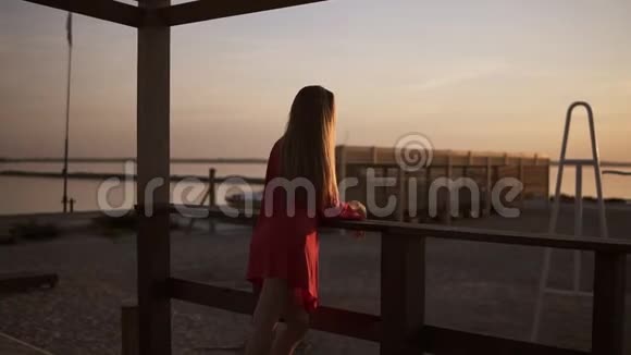 早晨大海或海洋日出的美丽镜头漂亮的长头发的金发女郎穿着红色连衣裙面向视频的预览图
