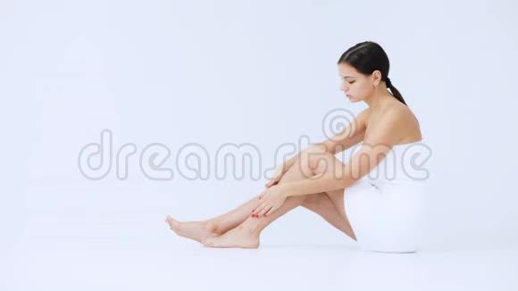 美丽的白种人年轻女子抚摸她的腿在她的腿上用清爽的奶油隔离在白色的背景上视频的预览图